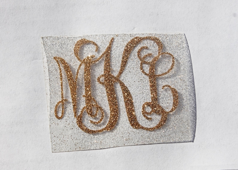 May include: A gold glitter monogram with the letters "MK" in a cursive font on a white background.