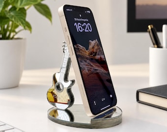 May include: A silver-colored metallic smartphone stand shaped like an acoustic guitar, featuring a reflective mirror finish on the guitar body and a circular base. A smartphone is resting against the stand, displaying the time 16:20 on its screen.