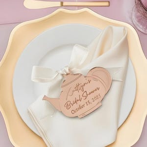 May include: A table setting with gold cutlery, a white plate, and a gold-rimmed charger. A white napkin is tied with a ribbon and holds a decorative teapot-shaped tag with text. Pink flowers and glassware are in the background.