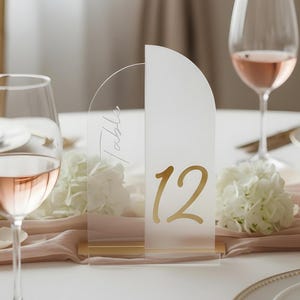 May include: A table setting with a clear acrylic table number 12 in gold script. The table is set with wine glasses filled with rose wine, white flowers, and a blush-colored runner. The setting is elegant and suitable for a wedding or special event.