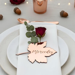 Peut inclure: Une table joliment dressée avec des couverts, des bougeoirs et des marque-places en forme de feuilles de couleur cuivre. Des assiettes et des serviettes blanches sont disposées avec des roses rouges et des feuilles d'automne. Le marque-place indique "Mercedes".