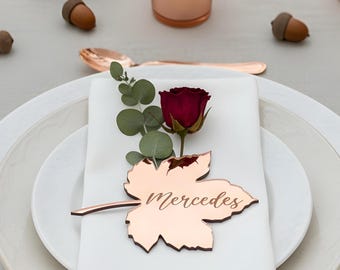 May include: A table setting featuring white plates, a white napkin, and a copper-colored leaf-shaped place card with the name Mercedes engraved on it. The place card is decorated with a single red rose and a sprig of eucalyptus, accompanied by copper cutlery.