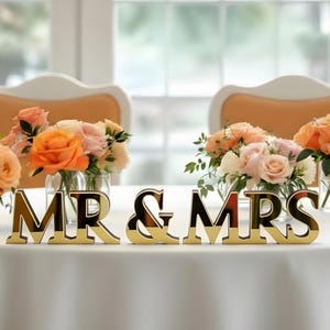 May include: A wedding reception table is set with floral arrangements in glass vases. The table features gold-colored "MR & MRS" lettering. The flowers are in shades of peach, orange, and cream, creating a romantic ambiance.