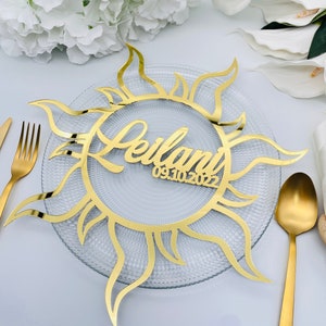 May include: A gold-coloured sun-shaped table decoration with the name "Leilani" and the date "09.10.2022" cut out in the centre. The sunburst design sits on a clear glass plate, with gold cutlery visible.