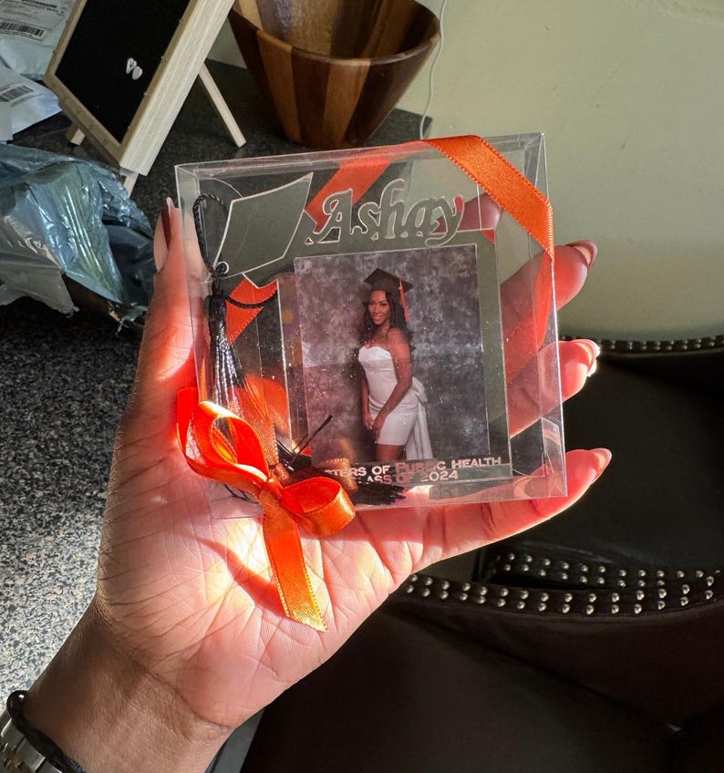 Puede incluir: Una caja de pl&aacute;stico transparente con una cinta rayada de color negro y naranja y un borla negra. Dentro de la caja hay un marco de fotos plateado con una foto de una mujer con un vestido blanco y un gorro de graduaci&oacute;n. El marco tiene el nombre "Ashay" grabado y el texto "Masters of Public Health Class of 2024".