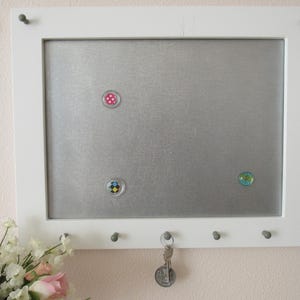 May include: White framed magnetic board with a silver metal surface. The board has three hooks on the bottom and three colorful magnets attached to the surface.