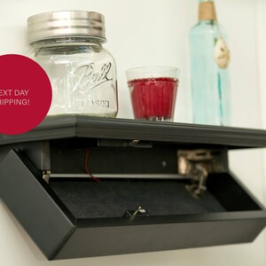 May include: A black shelf with a hidden compartment that opens to reveal a storage space. The shelf has a glass jar, a glass of red liquid, and a clear glass bottle on top.