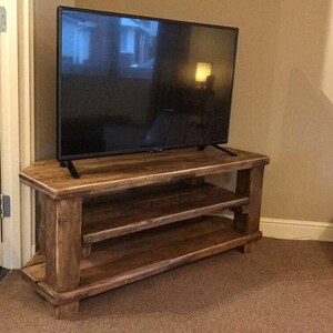 Op de afbeelding: Een rustieke houten tv-standaard met twee planken en een flatscreen televisie. De standaard is gemaakt van lichtbruin hout en heeft een eenvoudig, stevig ontwerp. De tv is zwart. De kamer heeft beige muren en een bruin tapijt. De standaard is ongeveer 120 cm breed.