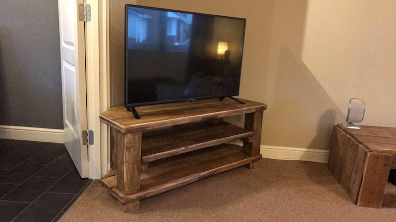 Op de afbeelding: Een rustieke houten tv-standaard met twee planken en een flatscreen televisie. De standaard is gemaakt van lichtbruin hout en heeft een eenvoudig, stevig ontwerp. De tv is zwart. De kamer heeft beige muren en een bruin tapijt. De standaard is ongeveer 120 cm breed.
