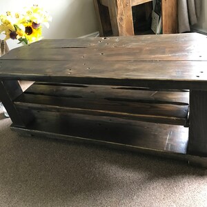Op de afbeelding: Een rustieke, donker gebeitste houten salontafel met een onderste plank. De tafel heeft een rechthoekig blad en stevige poten, met zichtbare houtnerf en knopen. De tafel is ongeveer 122 cm lang, 61 cm breed en 46 cm hoog.