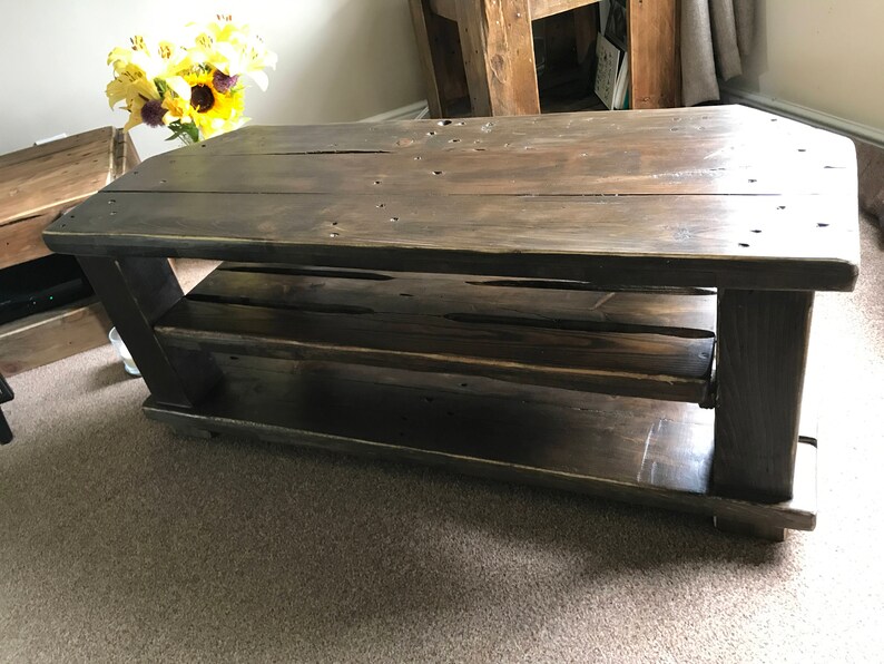 Op de afbeelding: Een rustieke, donker gebeitste houten salontafel met een onderste plank. De tafel heeft een rechthoekig blad en stevige poten, met zichtbare houtnerf en knopen. De tafel is ongeveer 122 cm lang, 61 cm breed en 46 cm hoog.