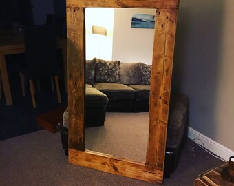 Op de afbeelding: Een grote, rechthoekige spiegel met een rustieke houten lijst. De lijst is gemaakt van lichtbruin, gerecycled hout met zichtbare knopen en onvolkomenheden. De spiegel weerspiegelt een kamer met een bank en kunst.