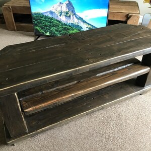 Op de afbeelding: Donker gebeitste houten tv-standaard met twee planken. De standaard heeft een uniek schuin ontwerp en is gemaakt van gerecycled hout. Het hout heeft zichtbare knopen en nerven, wat bijdraagt aan de rustieke charme. De standaard is ongeveer 122 cm lang.