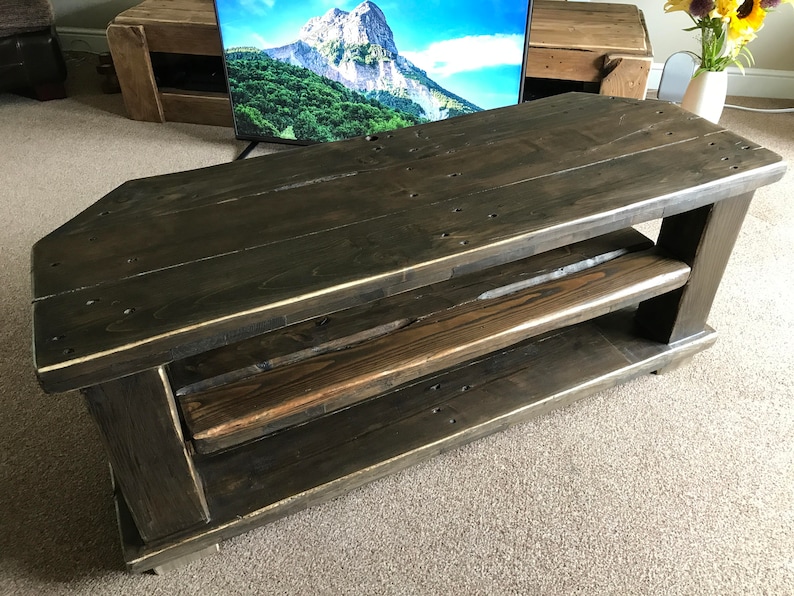 Op de afbeelding: Donker gebeitste houten tv-standaard met twee planken. De standaard heeft een uniek schuin ontwerp en is gemaakt van gerecycled hout. Het hout heeft zichtbare knopen en nerven, wat bijdraagt aan de rustieke charme. De standaard is ongeveer 122 cm lang.