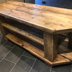 Op de afbeelding: Een rustieke houten tv-standaard met twee planken. De standaard is gemaakt van lichtbruin hout en heeft een natuurlijke afwerking. De standaard is ongeveer 122 cm lang, 46 cm breed en 51 cm hoog.