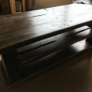 Op de afbeelding: Een donker gebeitste houten salontafel met twee planken. De tafel is rechthoekig met een lattenbovenkant en is gemaakt van dikke houten planken. De tafel is ongeveer 122 cm lang, 51 cm breed en 46 cm hoog.