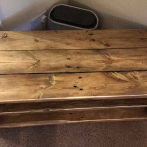Op de afbeelding: Een rustieke houten hoek-tv-standaard met een natuurlijke afwerking. De standaard heeft een lattenbovenkant en twee planken. Het hout vertoont knopen en nerven. Ontworpen om in een hoek te passen. Afmetingen: ongeveer 120 cm lang, 40 cm hoog en 40 cm diep.