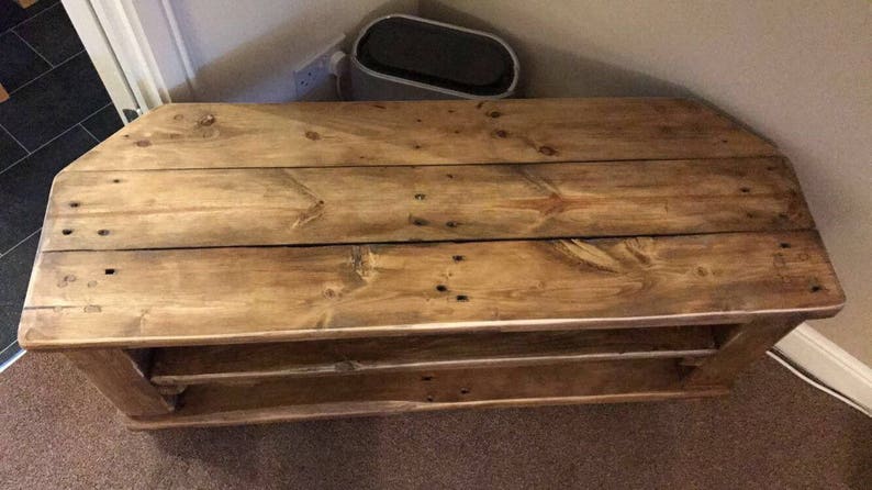 Op de afbeelding: Een rustieke houten hoek-tv-standaard met een natuurlijke afwerking. De standaard heeft een lattenbovenkant en twee planken. Het hout vertoont knopen en nerven. Ontworpen om in een hoek te passen. Afmetingen: ongeveer 120 cm lang, 40 cm hoog en 40 cm diep.