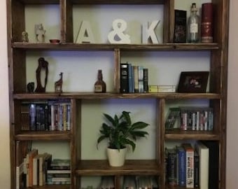 Op de afbeelding: Een grote, rustieke houten boekenkast met meerdere compartimenten. De planken zijn gevuld met boeken, decoratieve items en een potplant. De letters "A & K" staan op een plank. De boekenkast is gebeitst in een warme bruine kleur.