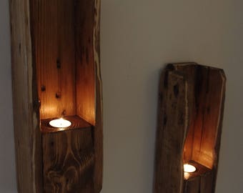 Op de afbeelding: Twee rustieke houten kandelaars aan de muur. Elke houder is een rechthoekig blok met een uitsparing voor een brandende waxinelichtkaars. Het hout heeft een natuurlijke, warme bruine tint.