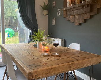 Op de afbeelding: Een rustieke houten eettafel met een lichtbruine afwerking, gecombineerd met witte stoelen en metalen haarspeldpoten. Twee wijnglazen en een brandende kaars staan op de tafel. Een decoratief houten plankje met kaarsen is aan de muur bevestigd.