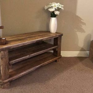 Op de afbeelding: Een rustieke houten tv-standaard met twee planken en een natuurlijke afwerking. Een witte vaas met witte bloemen staat erop. Een houten kandelaar met vijf kaarsruimtes staat links.