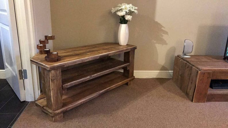 Op de afbeelding: Een rustieke houten tv-standaard met twee planken en een natuurlijke afwerking. Een witte vaas met witte bloemen staat erop. Een houten kandelaar met vijf kaarsruimtes staat links.