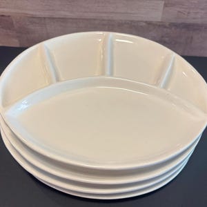 May include: A stack of four off-white, divided serving dishes. Each dish has three sections, with a rounded oval shape. The plates are stacked on a dark surface, with a light-colored wooden background.