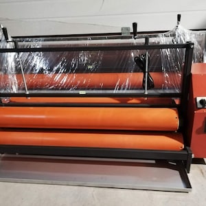 Peut inclure: Une machine industrielle noire avec deux rouleaux orange et un panneau de commande rouge. La machine est recouverte d'une pellicule plastique transparente.