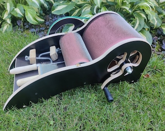 May include: A wooden wool carding machine with a black base and a crank handle. The machine has two rollers with pink bristles and a set of tools for carding wool.