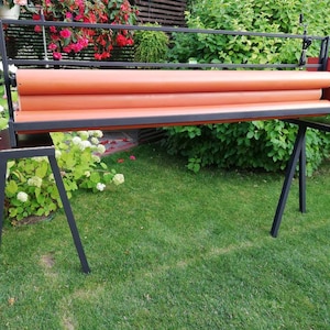 Op de afbeelding: Een zwarte metalen rolmachine met oranje stoffen rollen, gemonteerd op een zwarte metalen standaard met drie poten. De machine staat buiten op een gazon.