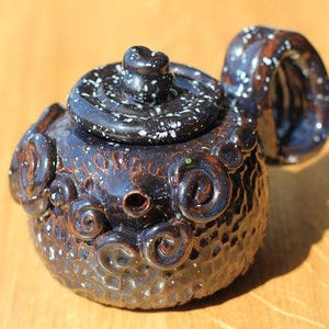 May include: A small, black and white ceramic teapot with a textured surface and a swirling design. The teapot has a spout and a lid.