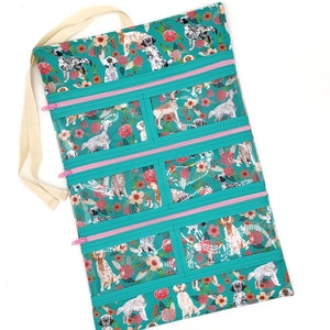 May include: Teal zippered pouch with clear vinyl windows, featuring a repeating pattern of white dogs and pink flowers. The pouch has a cream-colored fabric handle.