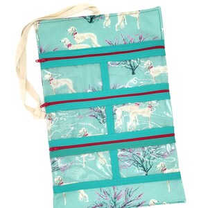 May include: Teal fabric jewelry roll with a white dog print and pink floral accents. The roll has five clear zippered compartments and a white canvas strap.