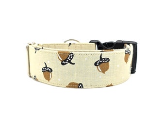 May include: A beige dog collar with a repeating pattern of brown acorns and black caps. The collar has a black plastic buckle and a silver metal D-ring for leash attachment. The fabric has a subtle, speckled texture.