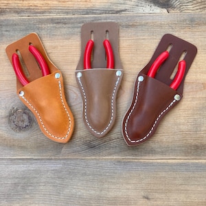 May include: Three leather tool holders with red-handled pliers inside. The holders are brown, tan, and dark brown.