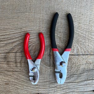 May include: Two slip-joint pliers with metallic silver heads and textured gripping jaws. The smaller pair features bright red rubberized handles, while the larger pair has black rubberized handles with red accents near the pivot point.