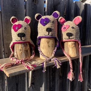 Pode incluir: Três chapéus de crochê em forma de rosto de urso feitos com fio rosa, roxo e branco. Cada chapéu tem um nariz em forma de coração com um botão preto para o olho. Os chapéus são exibidos em uma prateleira de madeira contra uma cerca de madeira.