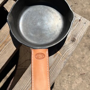 May include: A cast iron skillet with a leather handle cover. The handle cover is tan leather with white stitching and has the text "Allen James" and "Texline, TX" embossed on it.