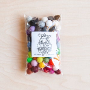 May include: A clear plastic bag filled with colorful felt balls. The bag has a label with a black and white illustration of a sheep and the text "Felt Ball Pack", "woolmob.com", and "@woolmob".