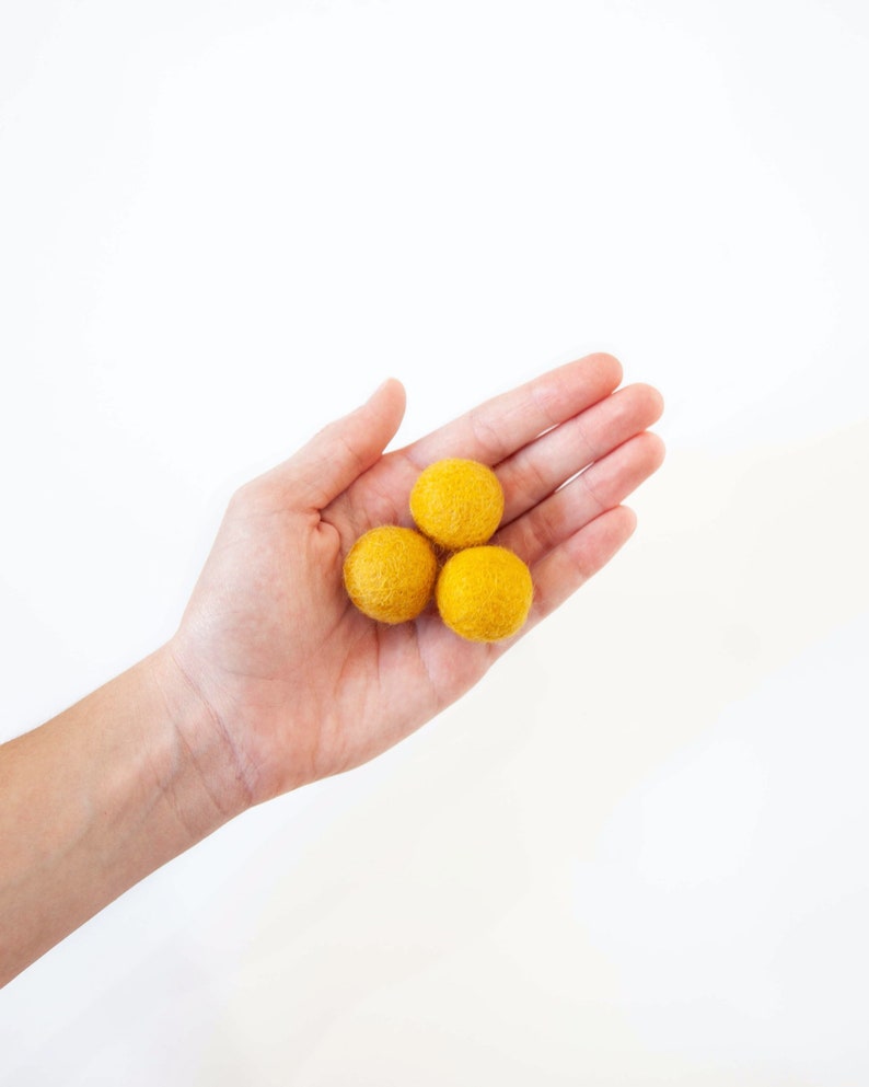 Hand showing the relative size of Wool Mob's Small felt balls. Each measures 2.5 cm or roughly 1 inch in diameter. Color shown is Sunrise or a rich, bright yellow.