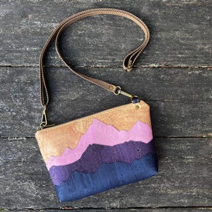 Op de afbeelding: Een kurken crossbodytas met een bergketenontwerp in tinten beige, roze, paars en marineblauw. De tas heeft een bruine riem en goudkleurige hardware. De tas ligt op een verweerd houten oppervlak.