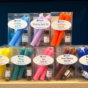 May include: A set of ten clear plastic boxes containing spell kits. Each box contains two candles and a small bottle of oil. The candles are in various colors, including blue, purple, orange, yellow, green, pink, brown, and blue. The labels on the boxes read "Cleansing Spell Kit", "Wishing Spell Kit", "Good Luck Spell Kit", "Head Opener Spell Kit", "Money Spell Kit", "Love Spell Kit", "Home Blessing Spell Kit", and "Protection Spell Kit".