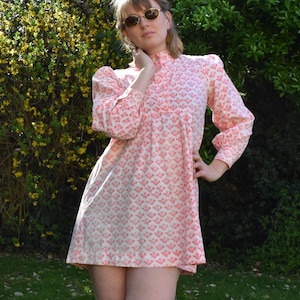 May include: A light pink, vintage-style dress with a floral pattern, high neckline, and long sleeves. The dress is worn with brown sandals. The image is set outdoors with a green lawn and foliage. The dress is approximately 80 cm in length.
