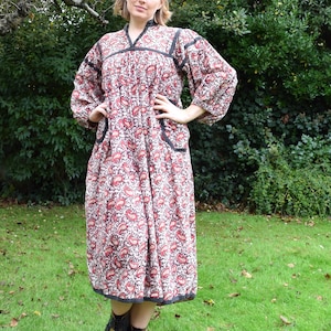 May include: A long, patterned dress with a red and white floral design. The dress has long sleeves, a V-neck, and black trim. The person is wearing black boots and standing on a grassy lawn.