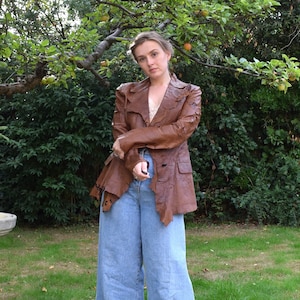 May include: A brown leather jacket with a distressed look, worn over a pair of light blue wide-leg jeans.