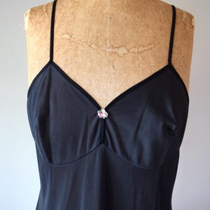 May include: A black slip dress with thin straps and a V-neckline. The neckline and straps are trimmed with a darker fabric. A small floral embellishment is centered at the bust. The dress is displayed on a vintage mannequin.