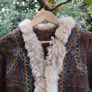 Könnte beinhalten: Eine braune Wildlederjacke mit einem weißen Schaffellfutter und bunten Stickereien. Die Jacke hängt an einem Holzbügel.