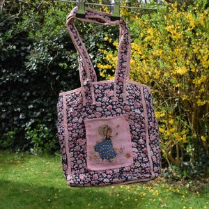 May include: A pink and blue floral tote bag with a pink trim and a patch featuring a girl in a blue dress and a pink hat.