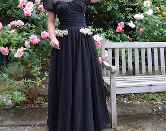 May include: A black tulle gown with a fitted bodice and a full skirt. The gown has a sheer overlay with a scoop neckline and short sleeves. The waist is cinched with a black sash and the skirt is adorned with a floral embellishment.